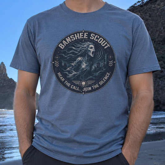 A man wearing a slate blue crewneck t-shirt with a circular 'Banshee Scout' graphic on the chest, set against a blurred beach and cliff background.