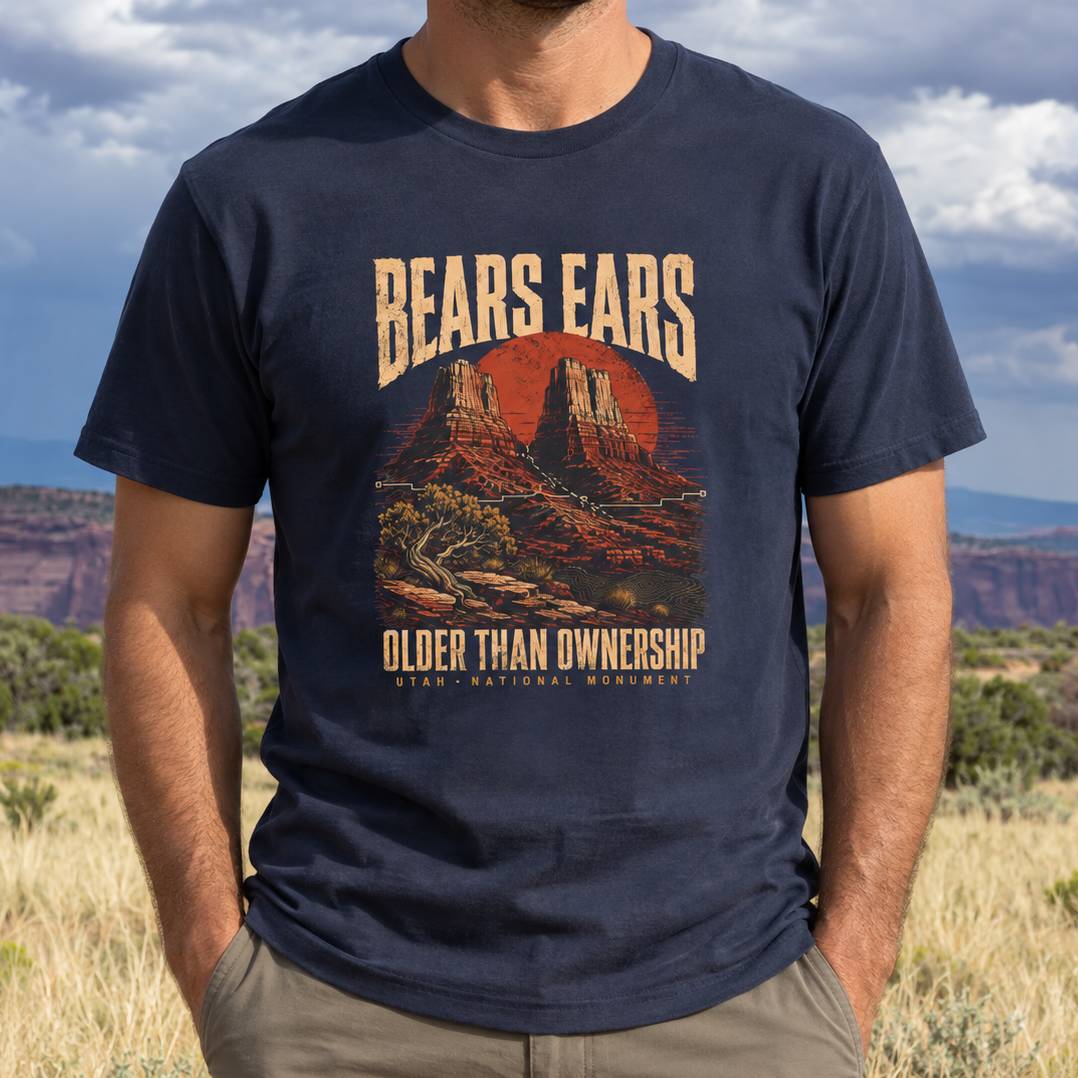 A man wearing a navy blue graphic t-shirt with a desert landscape print that reads 'BEARS EARS' and 'OLDER THAN OWNERSHIP'. The background shows a blurred outdoor desert landscape under a cloudy sky.