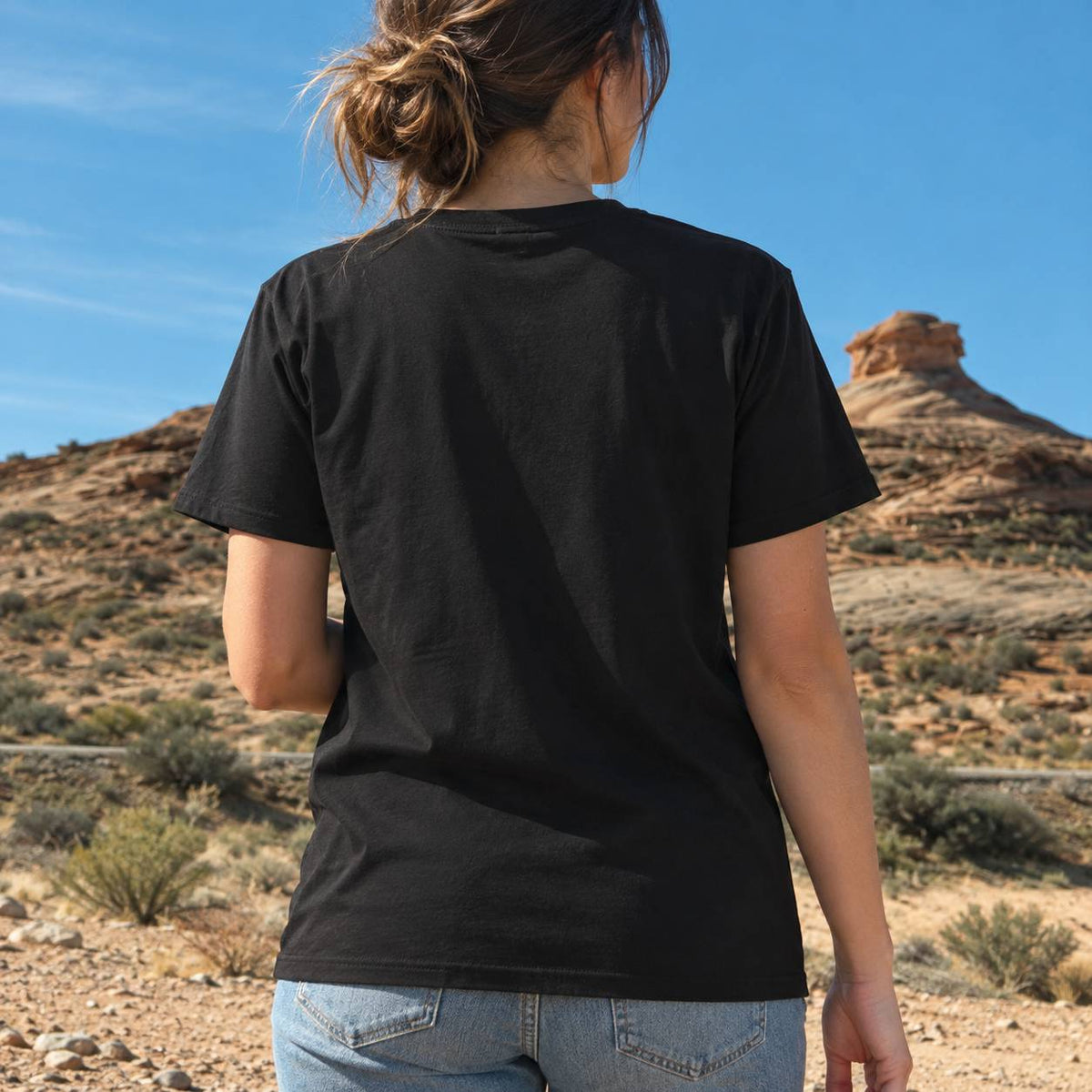 A woman viewed from behind wearing a plain black short-sleeve t-shirt and light blue jeans, standing in a desert landscape with rocky hills and a blue sky.
