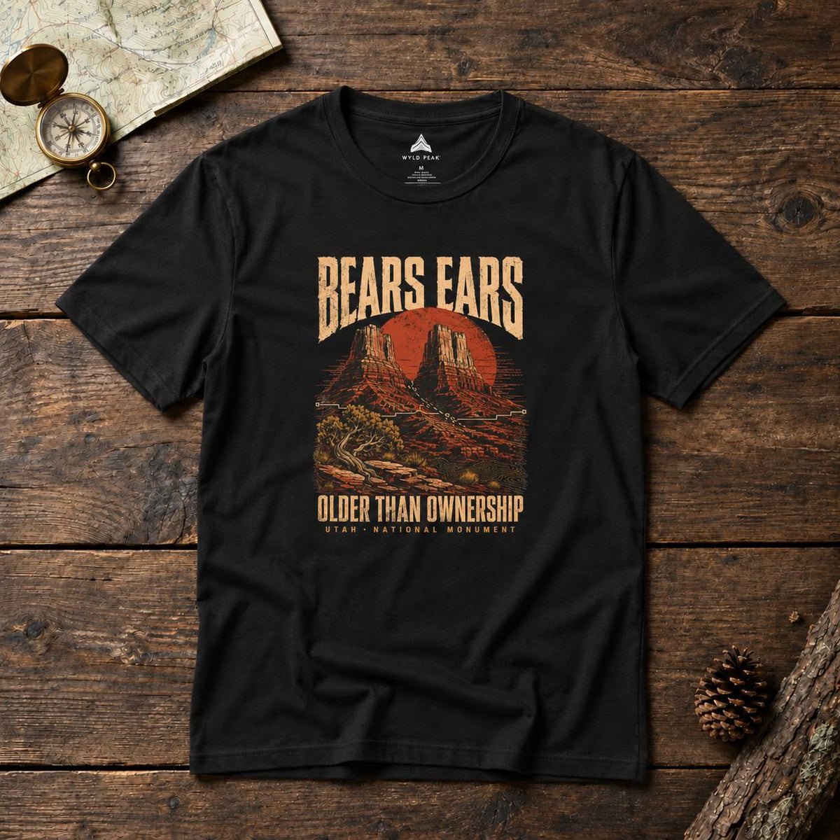 A black graphic t-shirt laid flat on a rustic wooden surface, featuring a vintage-style desert landscape print with the text 'BEARS EARS' and 'OLDER THAN OWNERSHIP'. The shirt is styled with a map, compass, and pinecone.