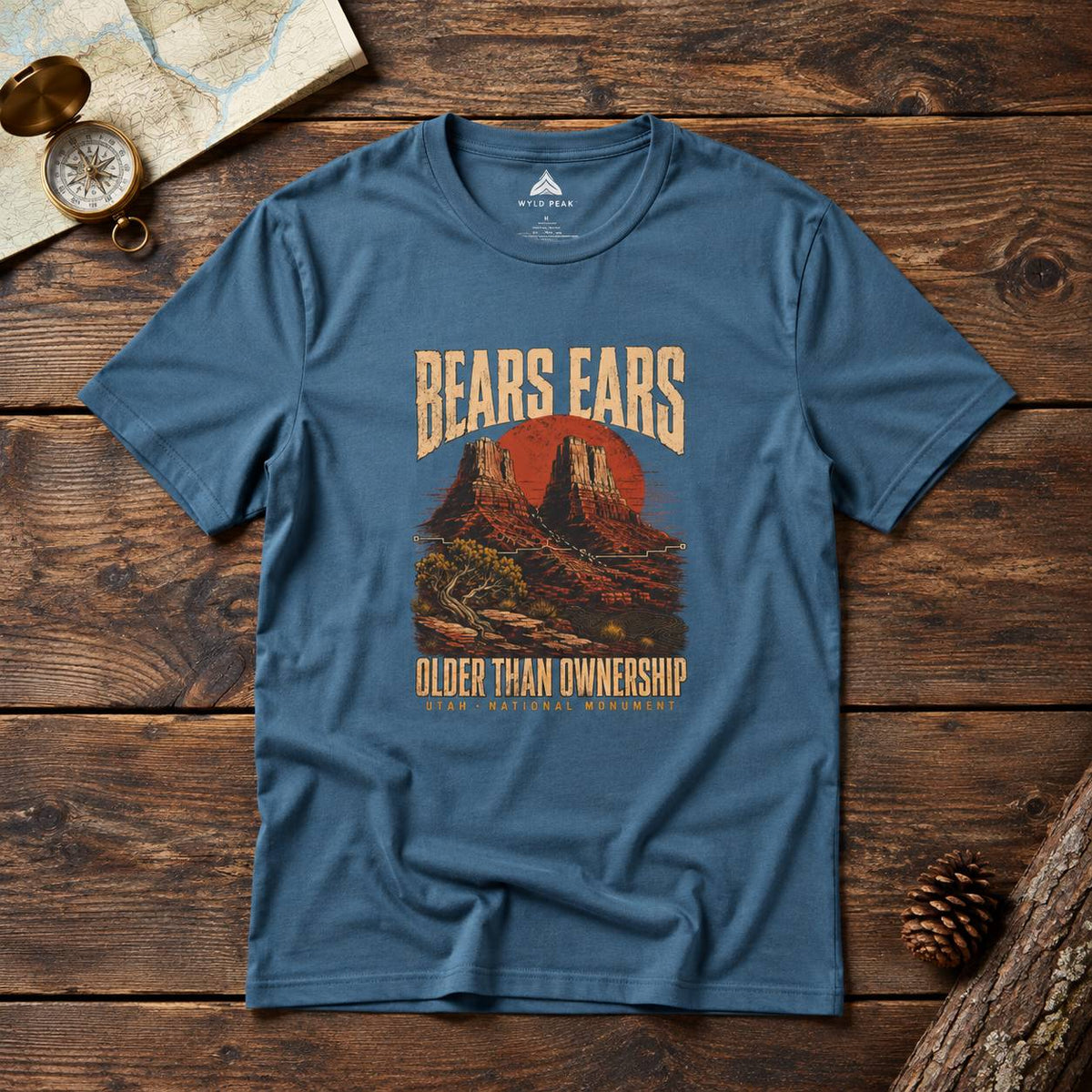 A blue crewneck t-shirt with a vintage-style graphic of a desert landscape and the text 'BEARS EARS OLDER THAN OWNERSHIP' laid flat on a rustic wooden background with a map, compass, and pinecone props.