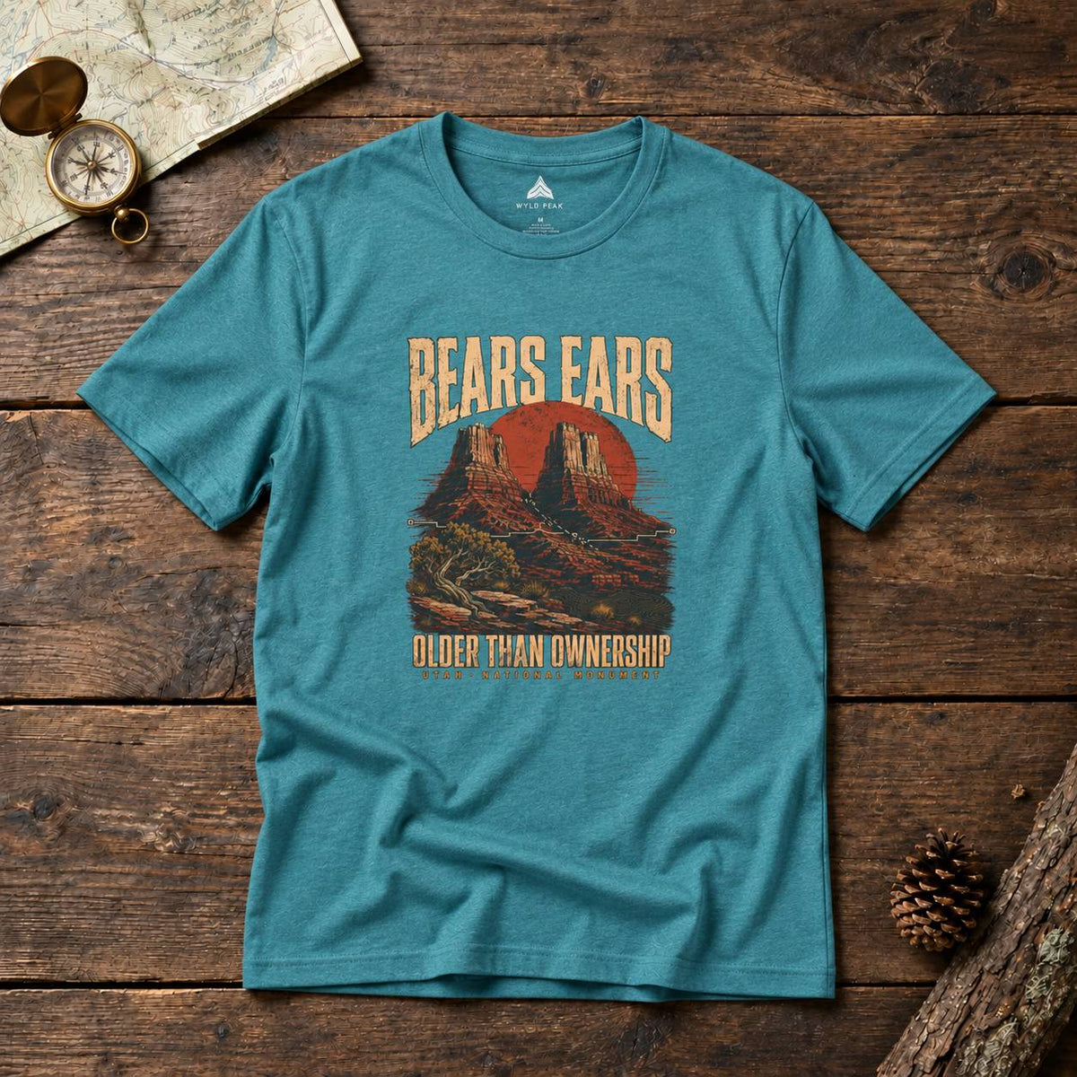 A teal short-sleeve graphic T-shirt laid flat on a rustic wooden surface, with a vintage-style desert landscape print and the words 'BEARS EARS' and 'OLDER THAN OWNERSHIP.' A map, compass, and pinecone are placed around it as styled props.