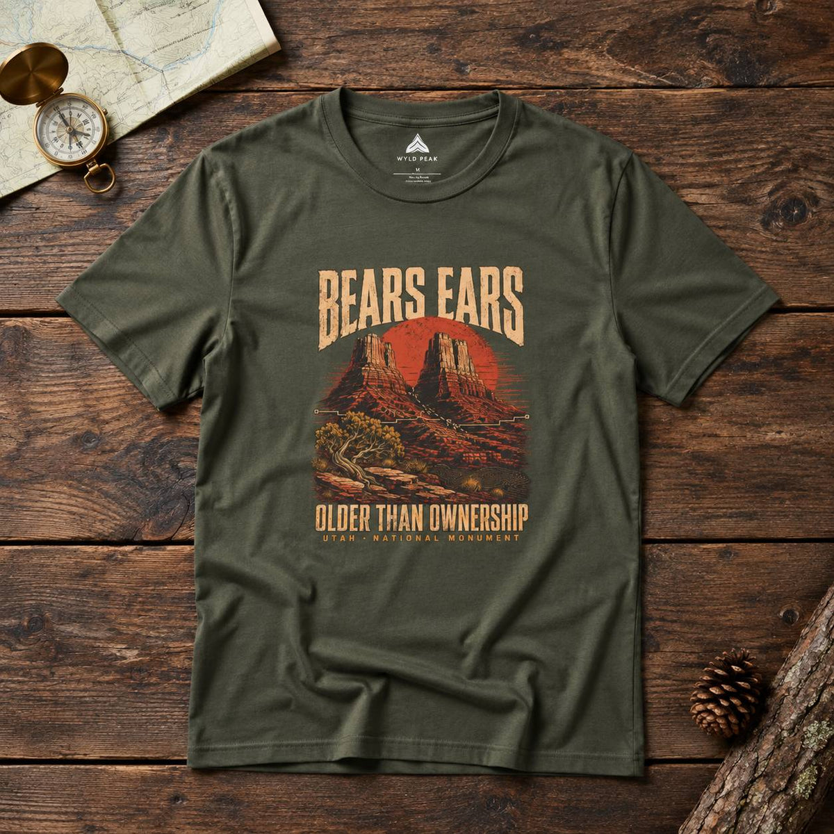 An olive green graphic t-shirt laid flat on a rustic wooden surface, surrounded by a vintage map, compass, pinecone, and wood log. The t-shirt features a retro-style graphic of a red sunset over desert mountains with the text 'BEARS EARS' and 'OLDER THAN OWNERSHIP' in beige.