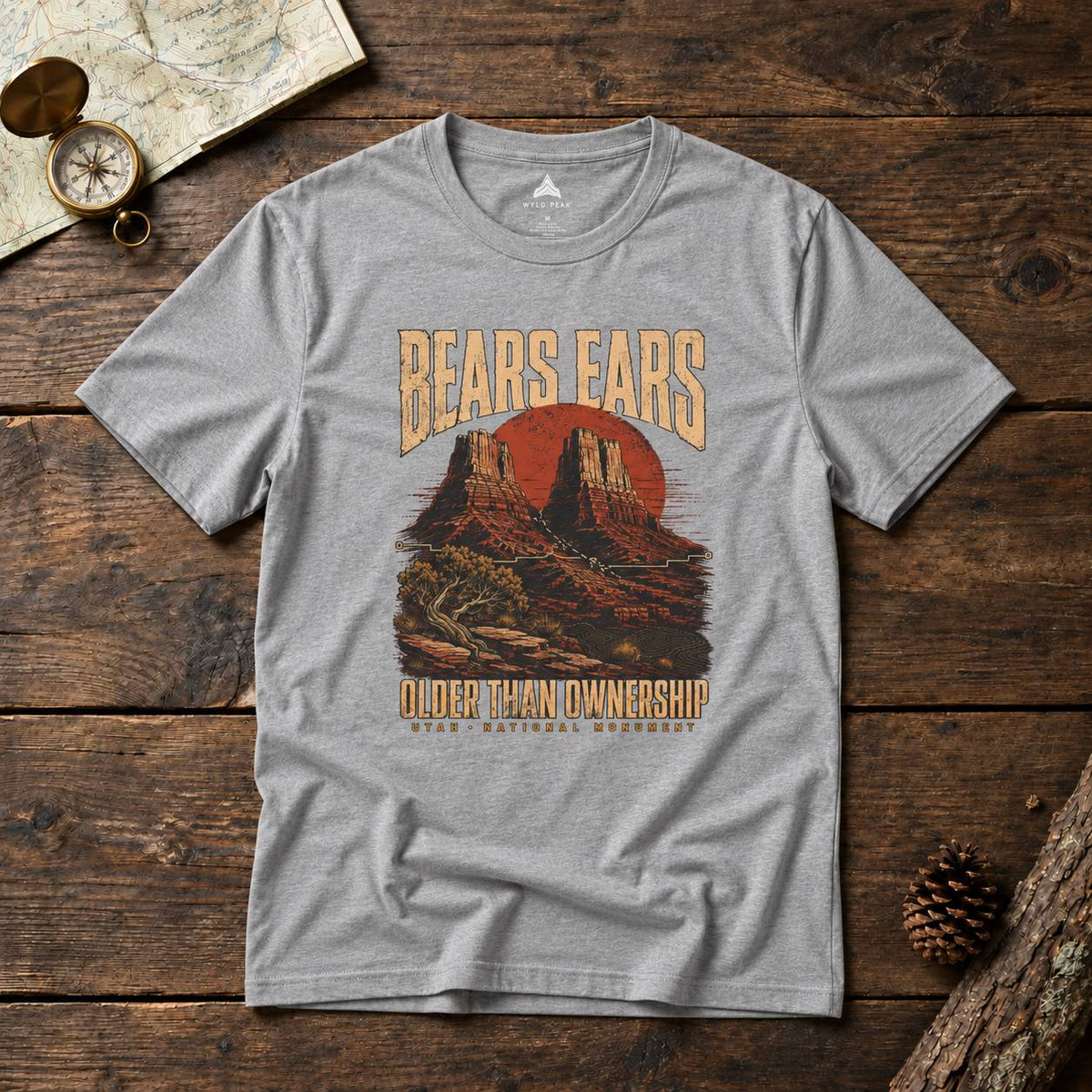 A heather gray graphic t-shirt laid flat on a rustic wooden surface. The shirt features a vintage-style desert landscape graphic with the text 'BEARS EARS' and 'OLDER THAN OWNERSHIP' with 'UTAH National Monument' printed below.