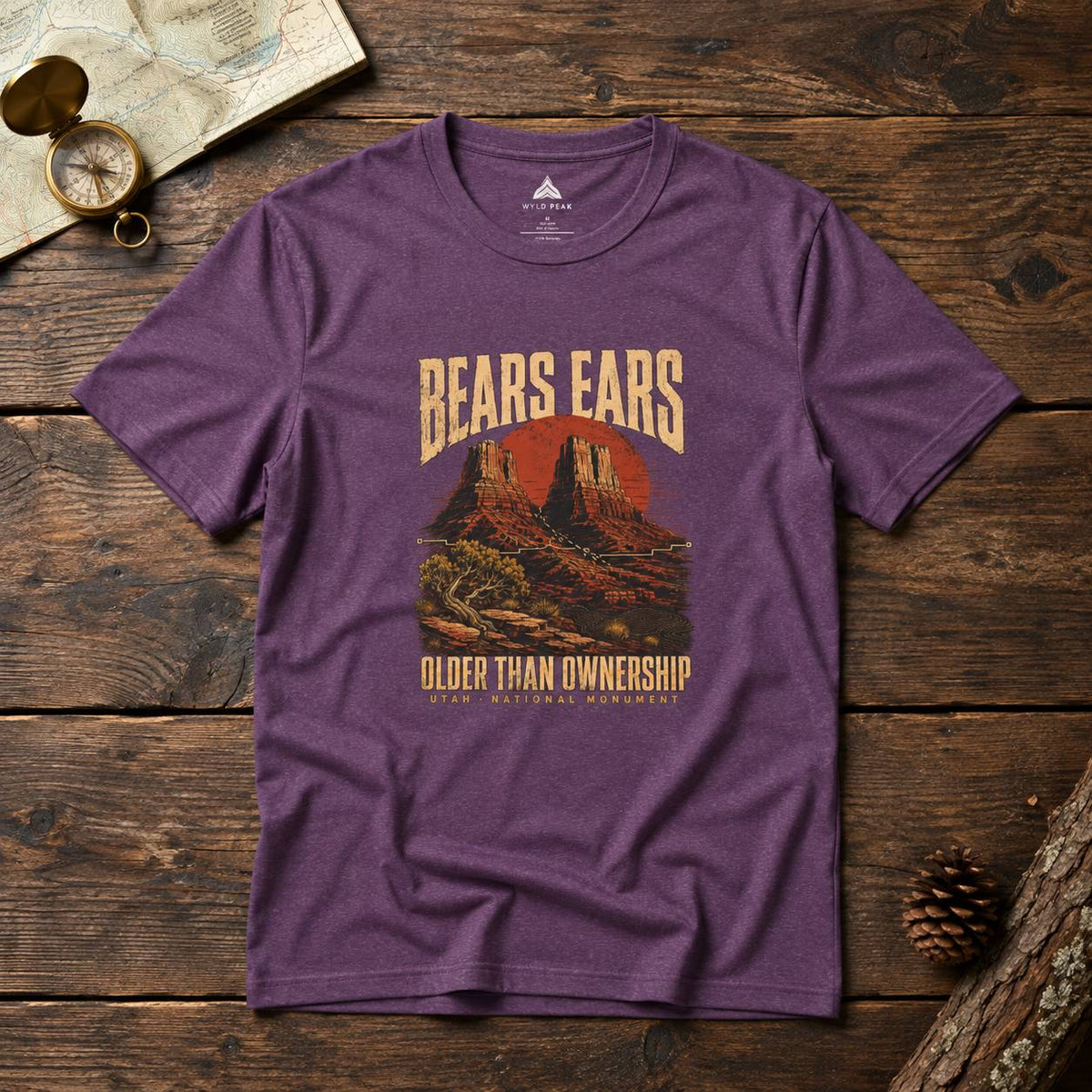 A heather purple graphic t-shirt laid flat on a rustic wooden surface, featuring a vintage-style red and beige desert landscape print with the text 'BEARS EARS' and 'OLDER THAN OWNERSHIP'.