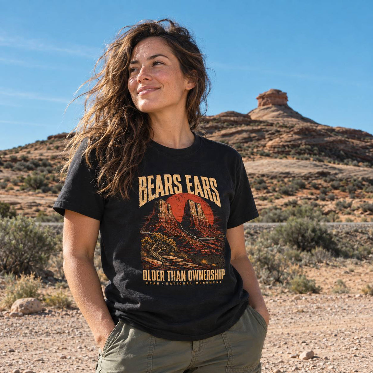 A woman wearing a black graphic t-shirt with a desert landscape print and the text 'BEARS EARS OLDER THAN OWNERSHIP' stands outdoors in a desert setting with a rock formation and blue sky in the background.