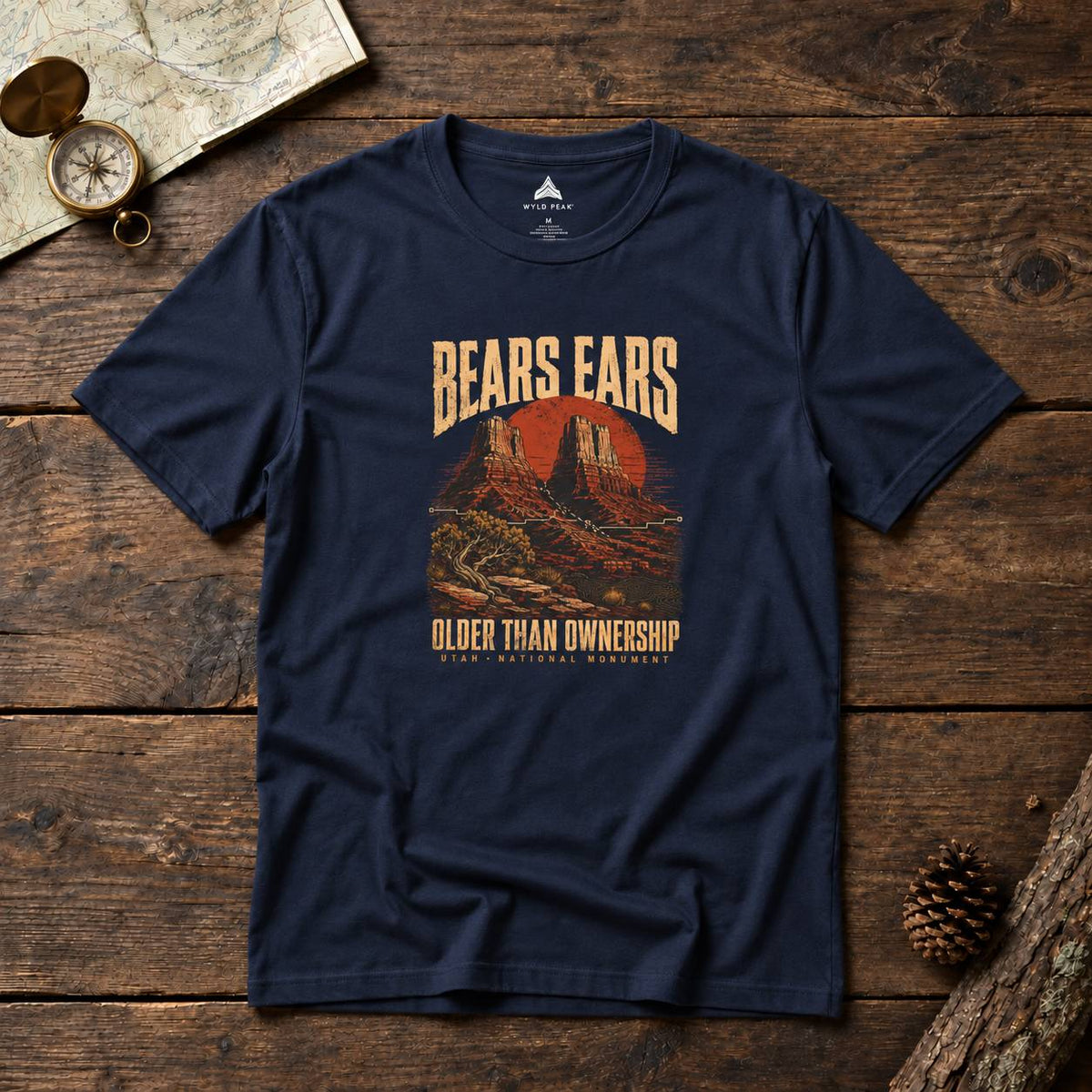 A navy blue graphic t-shirt laid flat on a rustic wooden surface, surrounded by vintage map props, a pinecone, and a small brass compass. The shirt features a retro-style desert landscape graphic with the text 'BEARS EARS' and 'OLDER THAN OWNERSHIP'.