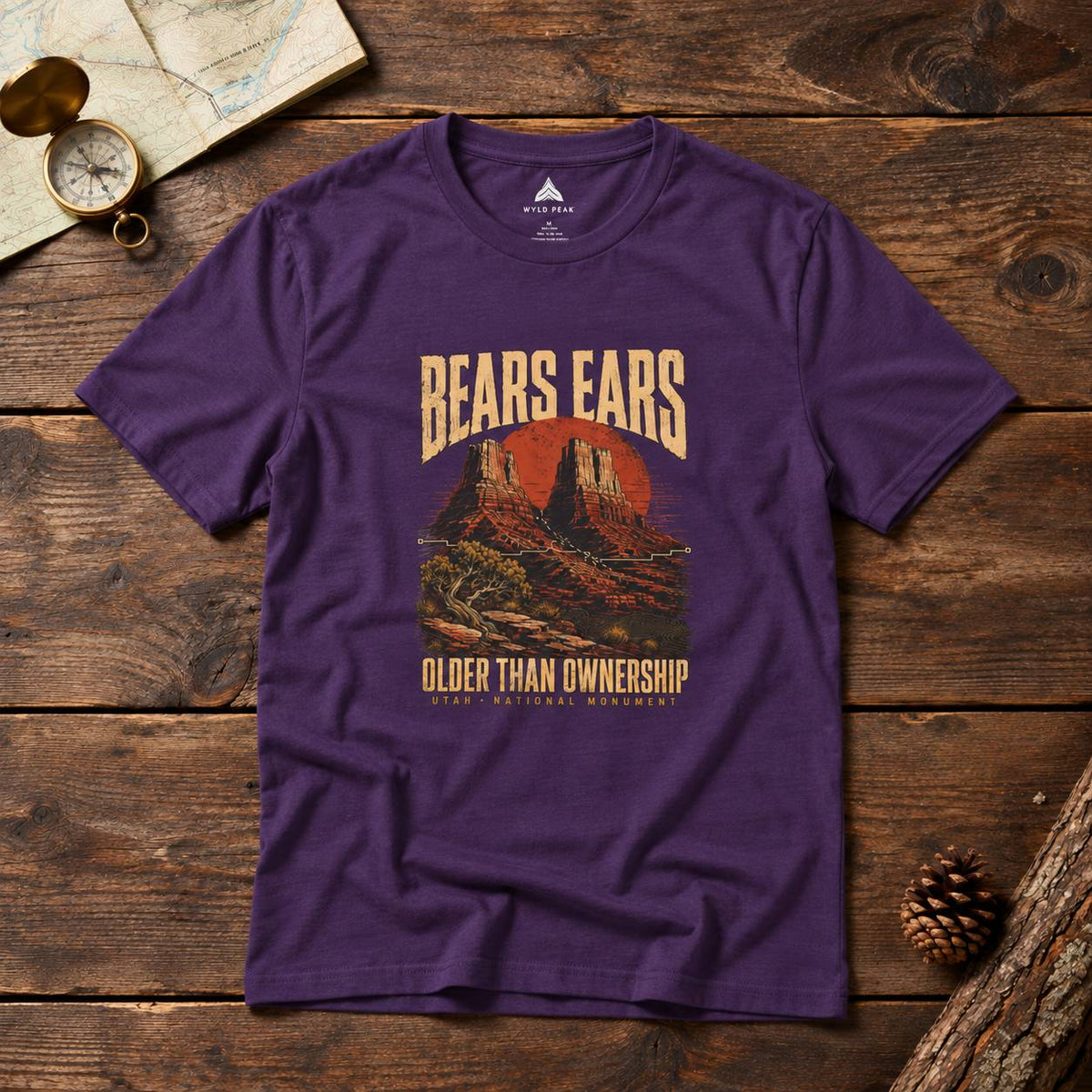 A purple short-sleeve graphic t-shirt laid flat on a rustic wooden surface, featuring a vintage-style red and beige desert landscape print with the text 'BEARS EARS' and 'OLDER THAN OWNERSHIP'.