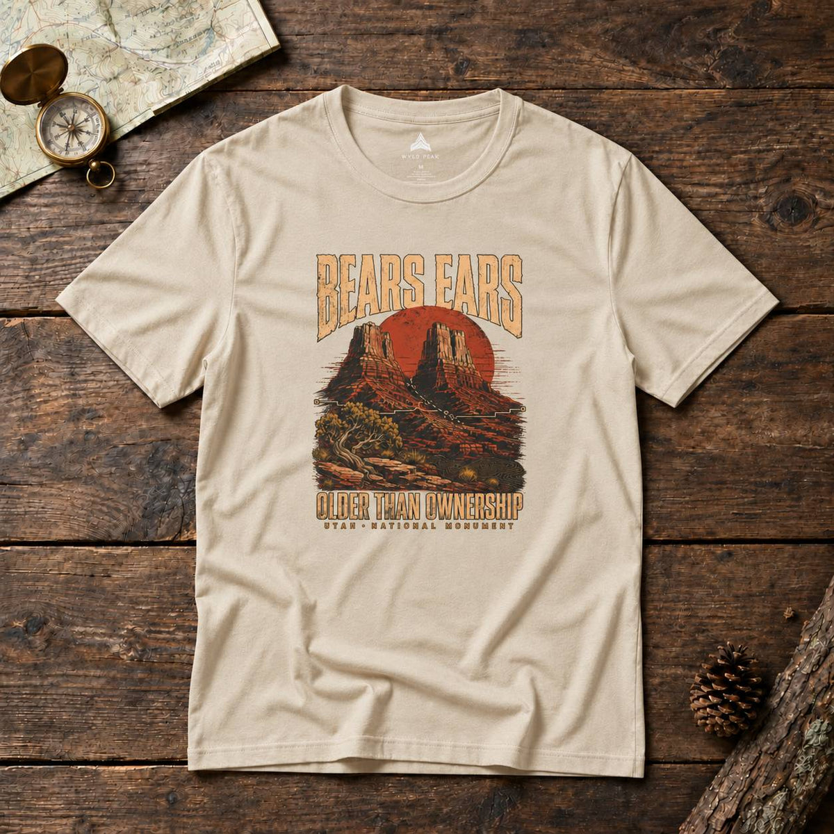A beige graphic t-shirt laid flat on a rustic dark wood background, featuring a desert landscape print with the text 'BEARS EARS' and 'OLDER THAN OWNERSHIP'.