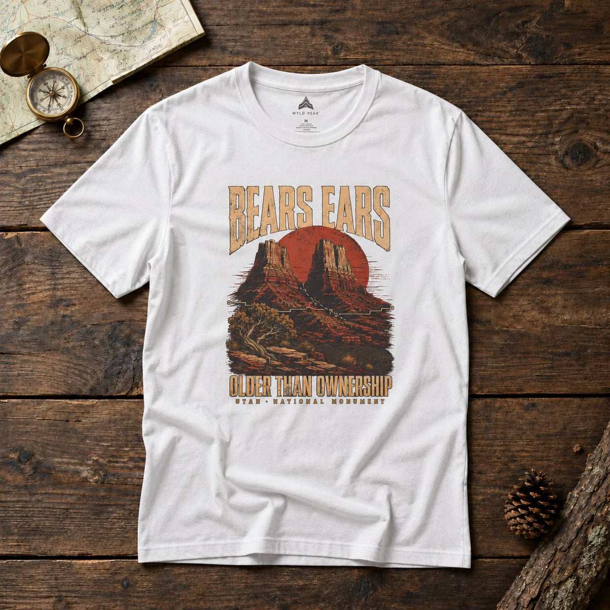 A white short-sleeve graphic T-shirt laid flat on a rustic dark wood surface, with a vintage-style desert mountain print and the text 'BEARS EARS' and 'OLDER THAN OWNERSHIP - HUAN NATIONAL MONUMENT.' Decorative props including a map, compass, and pinecone surround the shirt.