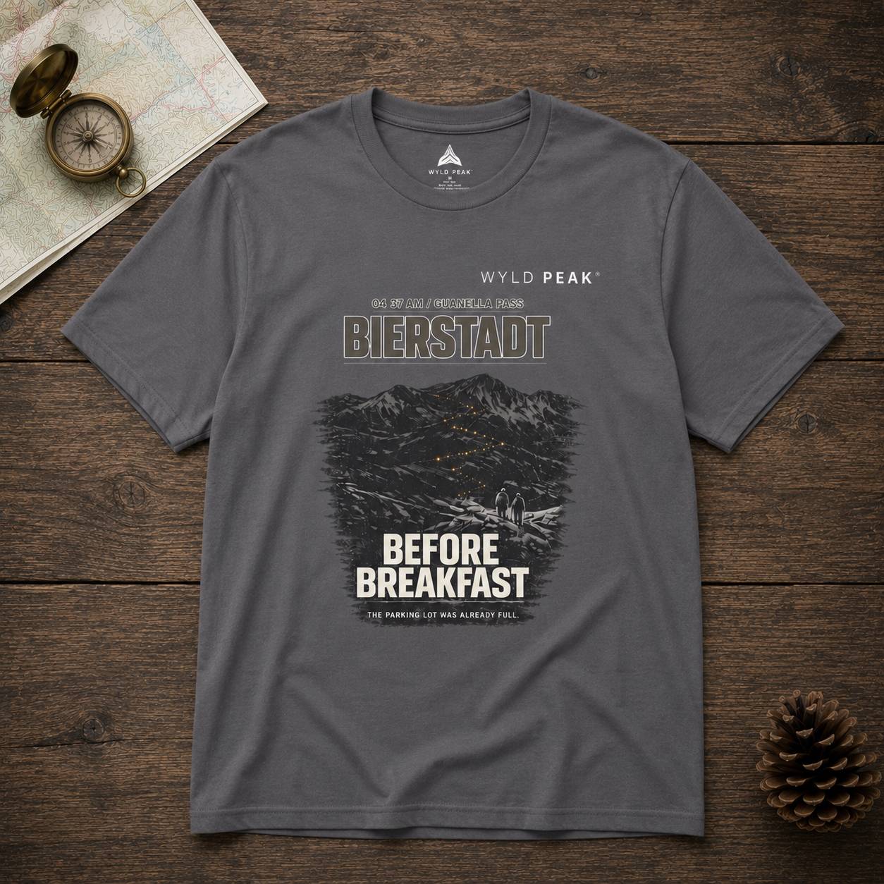 Gray t-shirt with mountain graphic and text on a wooden surface with map and compass.