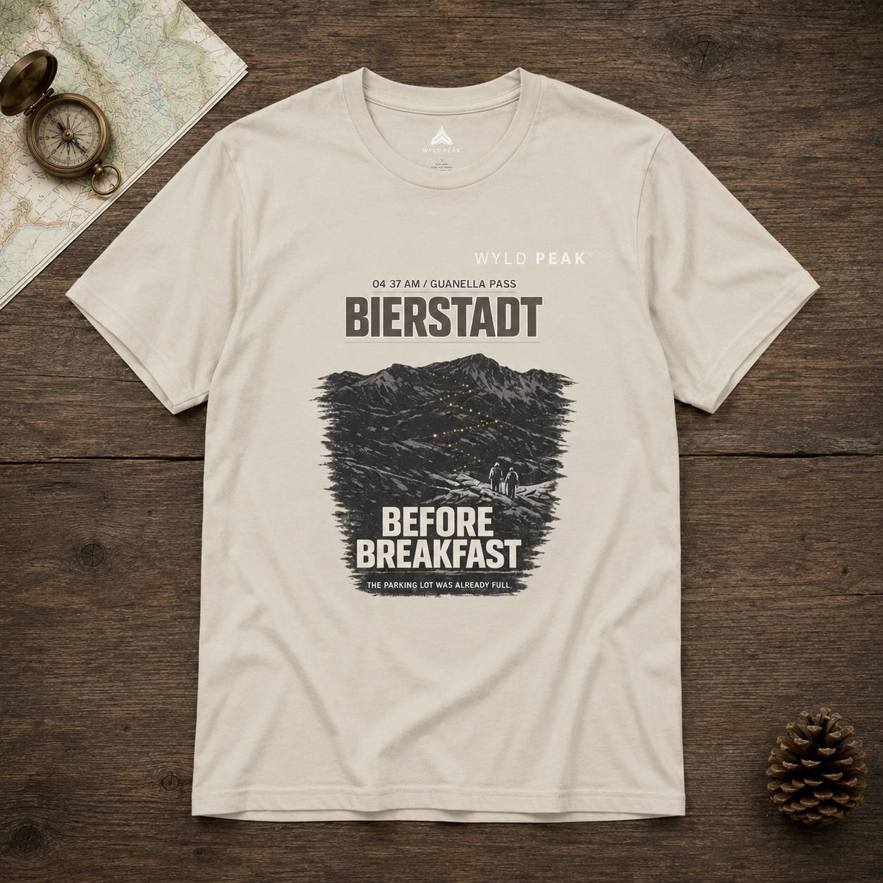Beige t-shirt with 'Bierstadt Before Breakfast' graphic on a wooden surface with map and compass.