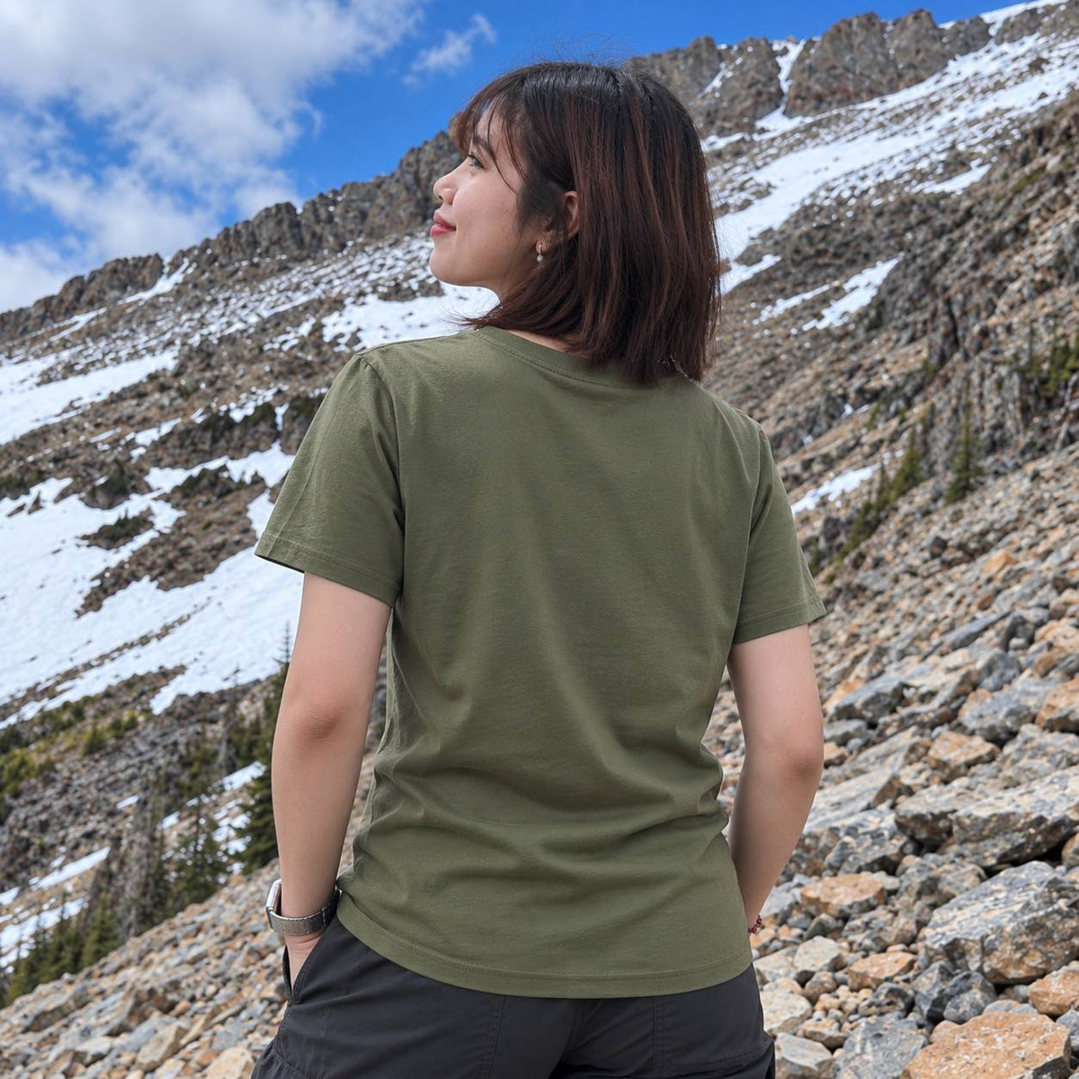 A woman wearing an olive green short-sleeve t-shirt and dark gray cargo pants, photographed from behind against a rocky mountain slope with patches of snow and a blue sky.