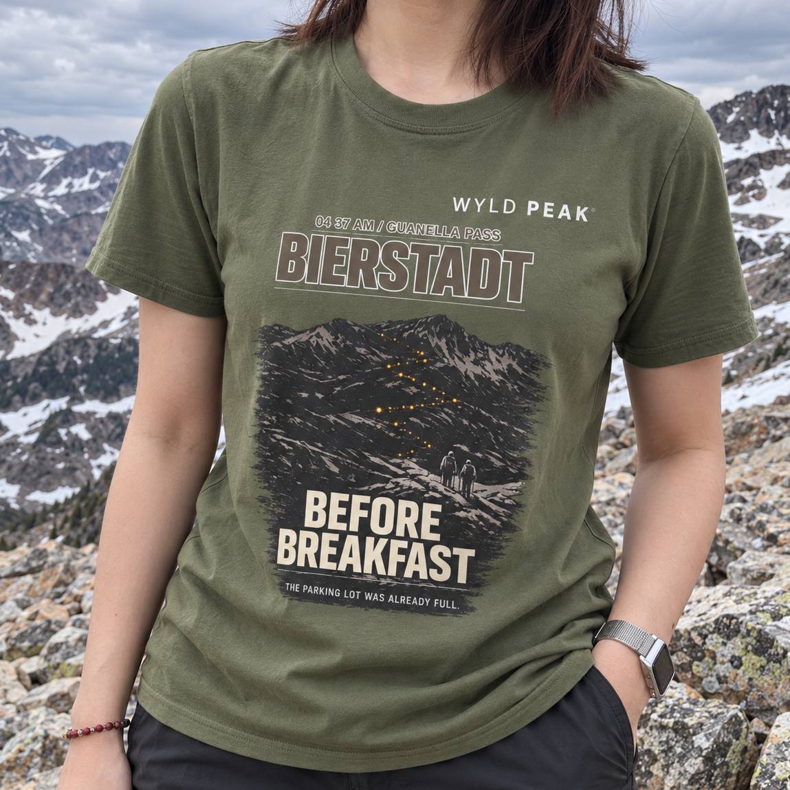 A person wearing an olive green graphic t-shirt with a mountain design and text reading 'WYLD PEAK', 'BIERSTADT', and 'BEFORE BREAKFAST', photographed outdoors against a rocky mountain landscape.