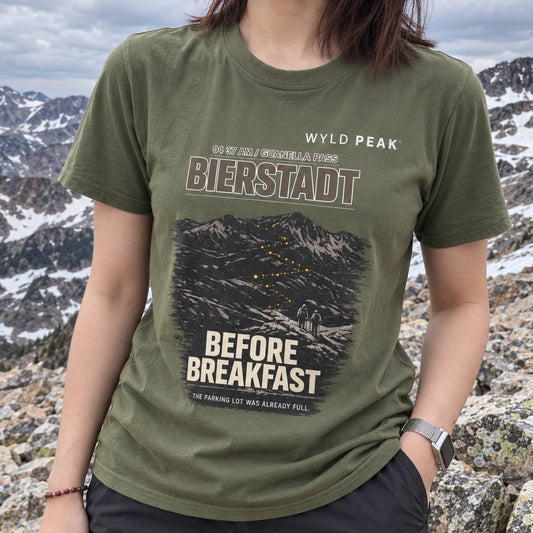A person wearing an olive green graphic t-shirt with a mountain design and text reading 'WYLD PEAK', 'BIERSTADT', and 'BEFORE BREAKFAST', photographed outdoors against a rocky mountain landscape.