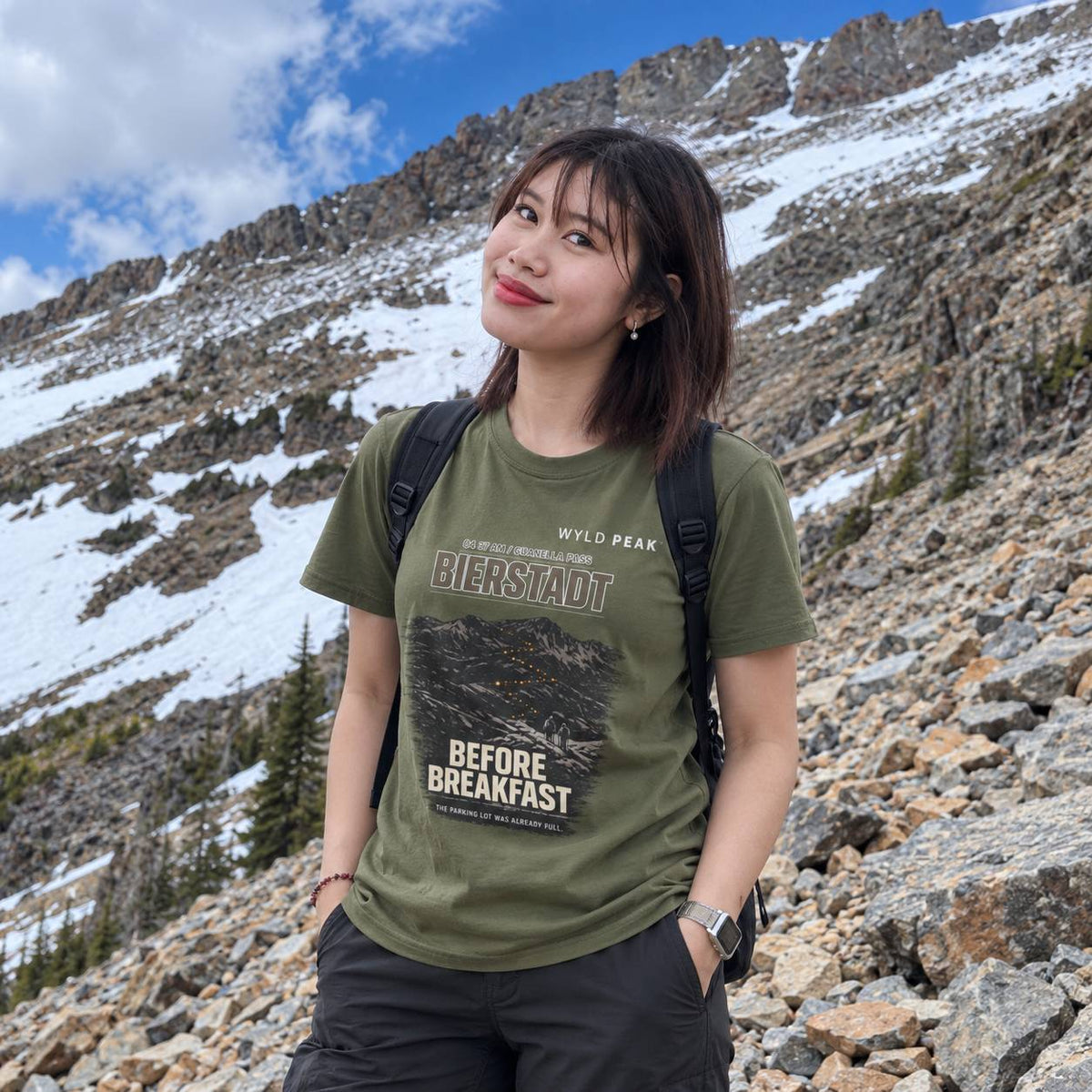 A woman wearing an olive green graphic t-shirt with 'BIERSTADT' and 'BEFORE BREAKFAST' printed on the front, posing outdoors on a rocky, snow-dusted mountain slope under a blue sky.
