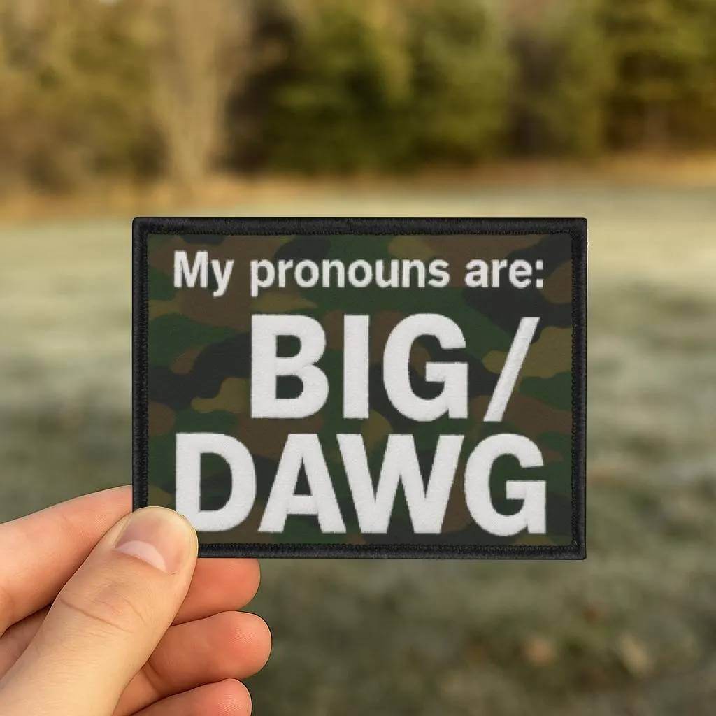 Person holding a patch with 'My pronouns are: BIG/DAWG' text against a blurred natural background