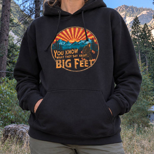 A person wearing a black pullover hoodie with a retro-style Bigfoot graphic on the chest, standing outdoors in a forested mountain setting.