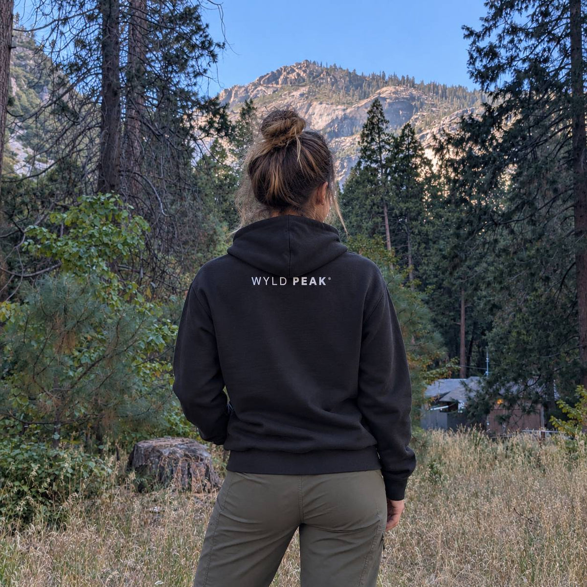 A woman viewed from behind wearing a black pullover hoodie with 'WYLD PEAK' printed in white on the back, standing in a grassy forest clearing with mountains and pine trees in the background.