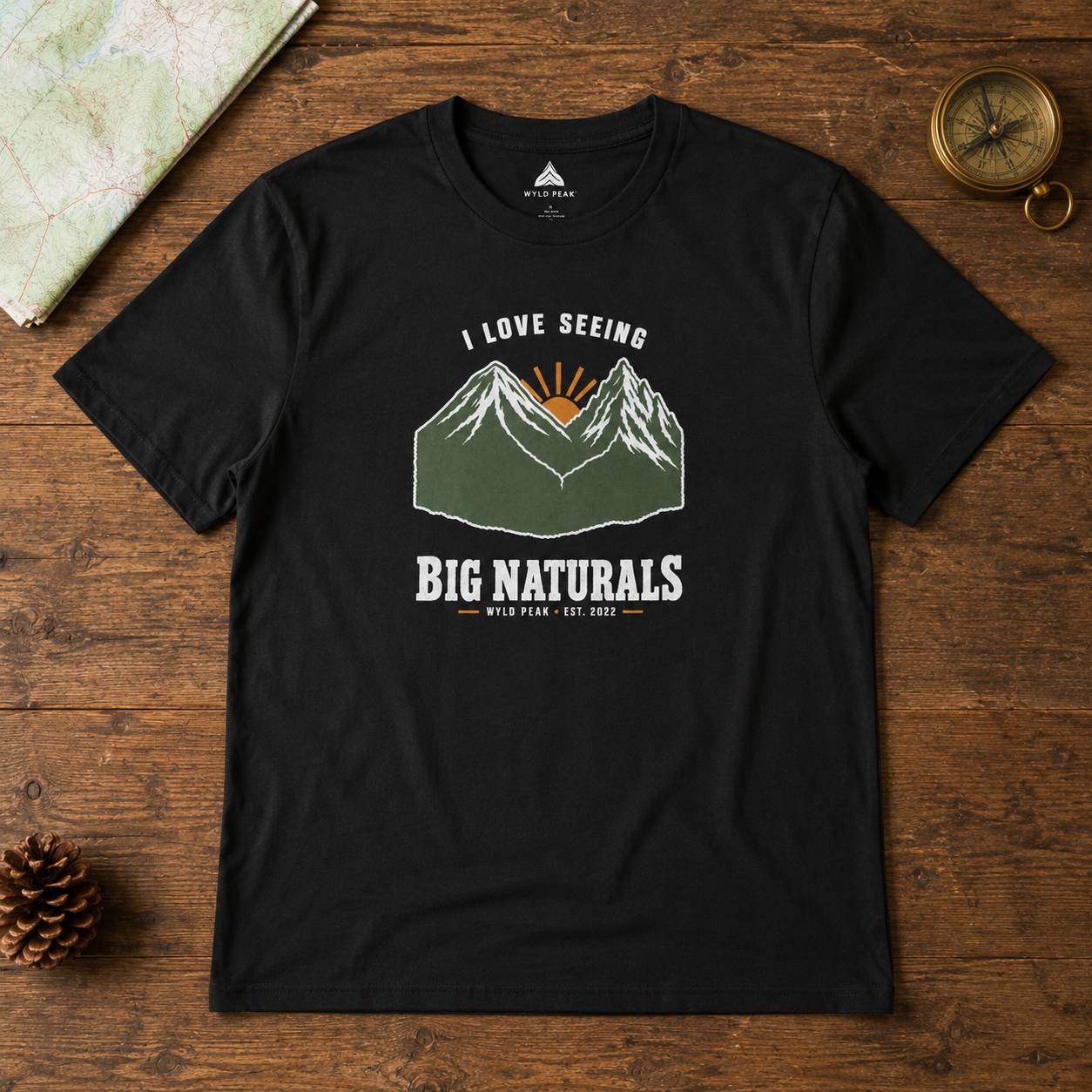 A black graphic t-shirt laid flat on a rustic wooden surface, featuring a green and white mountain design with a yellow sun and the text 'I LOVE SEEING BIG NATURALS'. The shirt is styled with outdoor props including a map, a pinecone, and a compass.