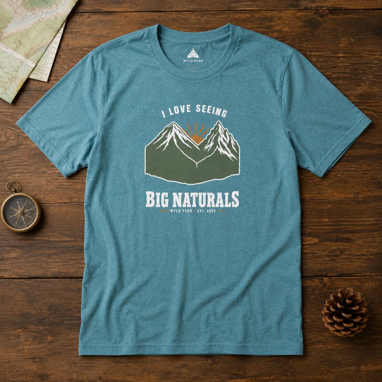 A heathered teal blue graphic t-shirt laid flat on a dark wood background, surrounded by a vintage compass, a map corner, and a pinecone. The t-shirt features a graphic of mountains with a rising sun and the text 'I LOVE SEEING BIG NATURALS WILD LIFE EST.