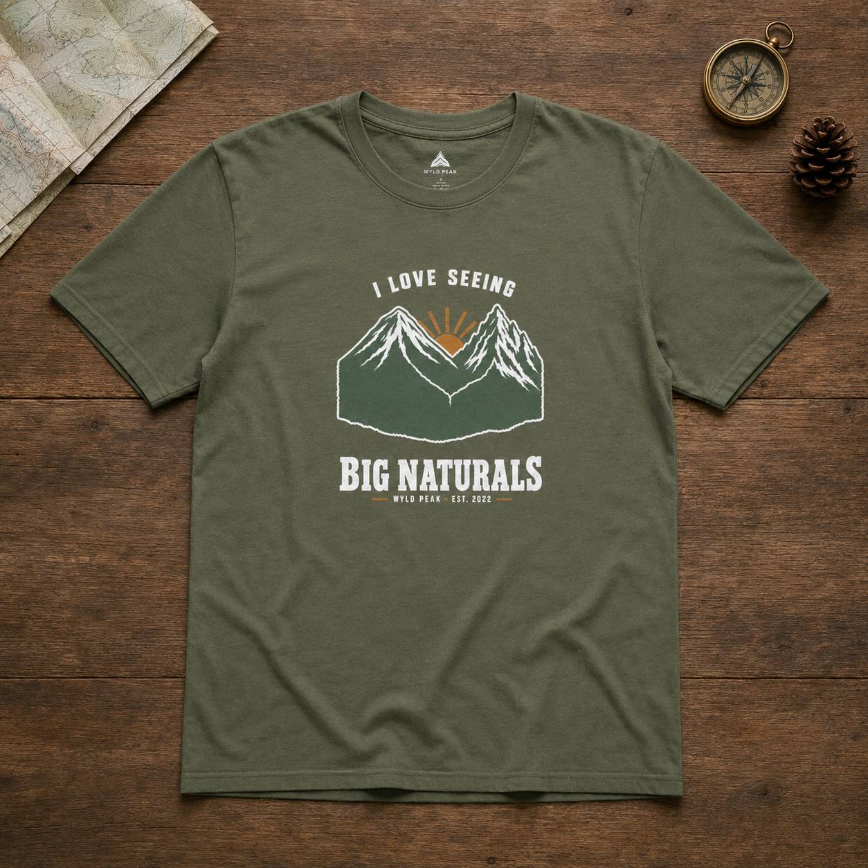 An olive green short-sleeve t-shirt with a mountain graphic and the text 'I LOVE SEEING BIG NATURALS' laid flat on a rustic wooden background with a compass, pinecone, and map as props.