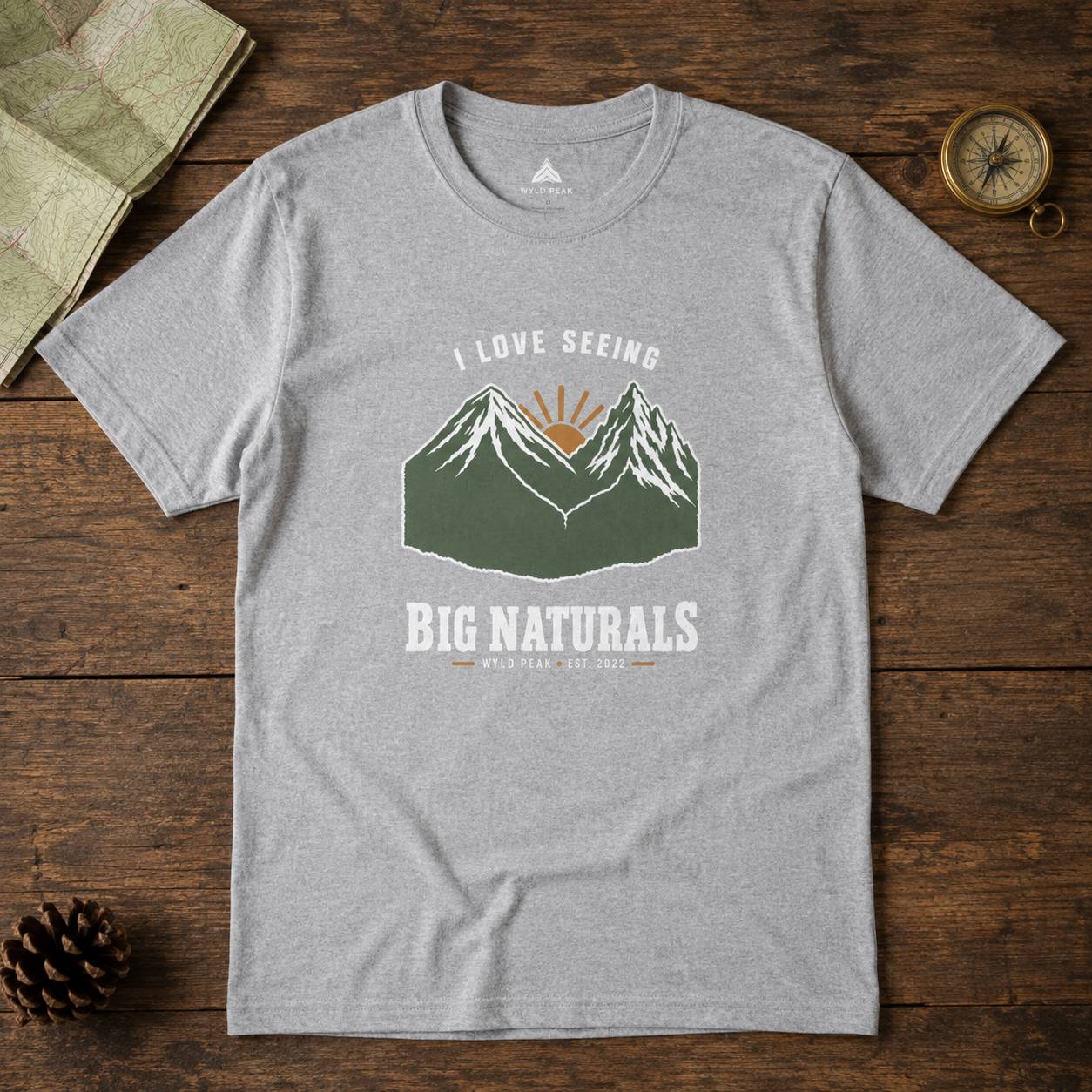 A heather gray short-sleeve t-shirt with a mountain graphic and the text 'I LOVE SEEING BIG NATURALS' laid flat on a rustic wooden surface with a map, pinecone, and compass as props.