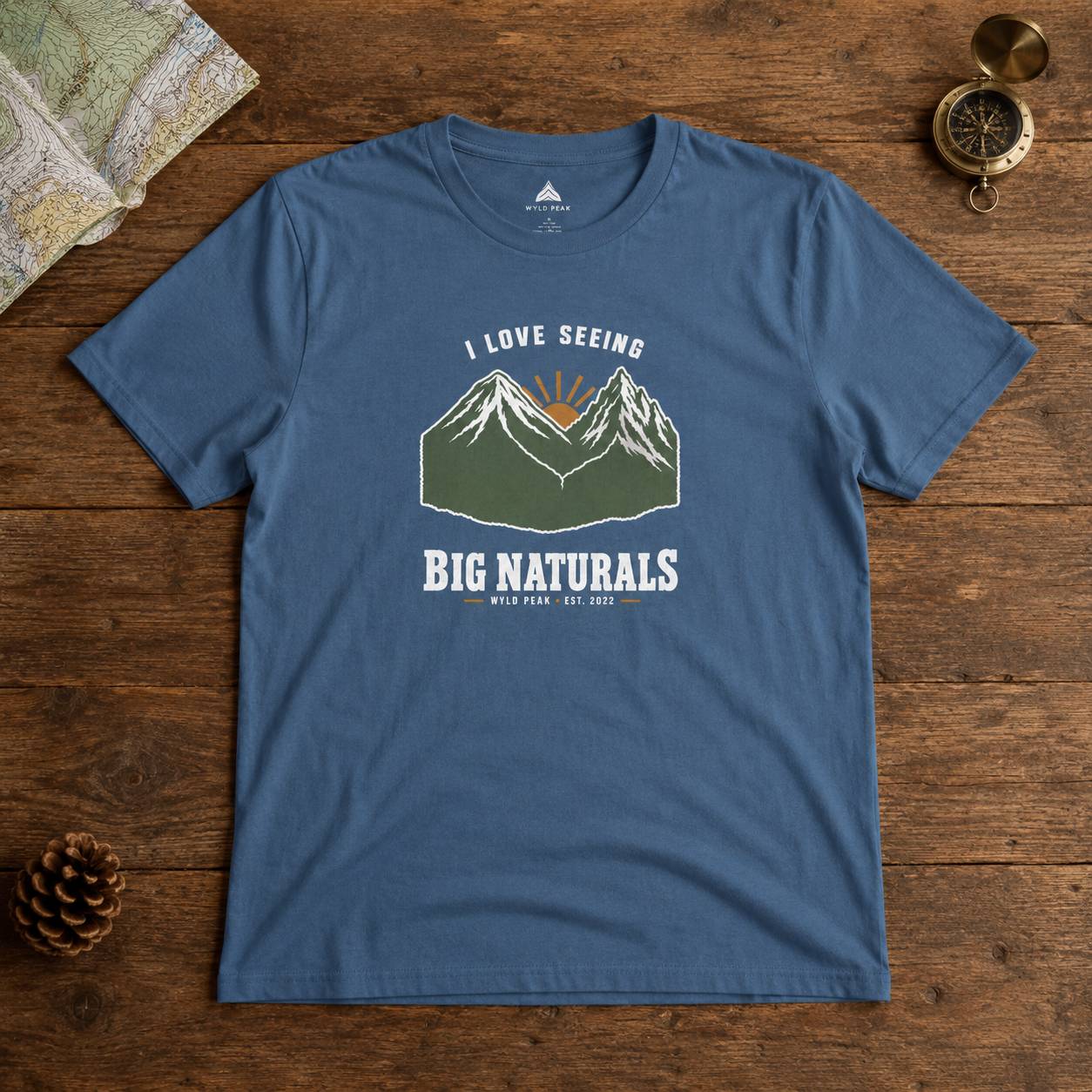 A blue crewneck t-shirt with a mountain graphic and the text 'I LOVE SEEING BIG NATURALS WILD PEAK EST. 2022' printed on the front, displayed flat on a rustic wooden background with a map, compass, and pinecone.