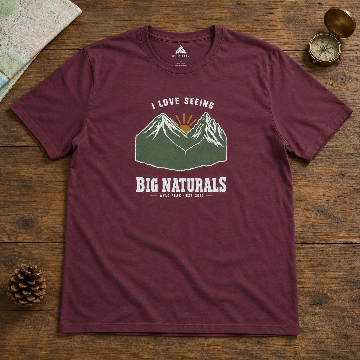 A maroon short-sleeve graphic t-shirt laid flat on a rustic wooden surface, surrounded by travel-themed props like a map, pinecone, compass, and brass lid. The t-shirt features a graphic of mountains with a sunrise and the text 'I LOVE SEEING BIG NATURALS WILD PEAK EST.