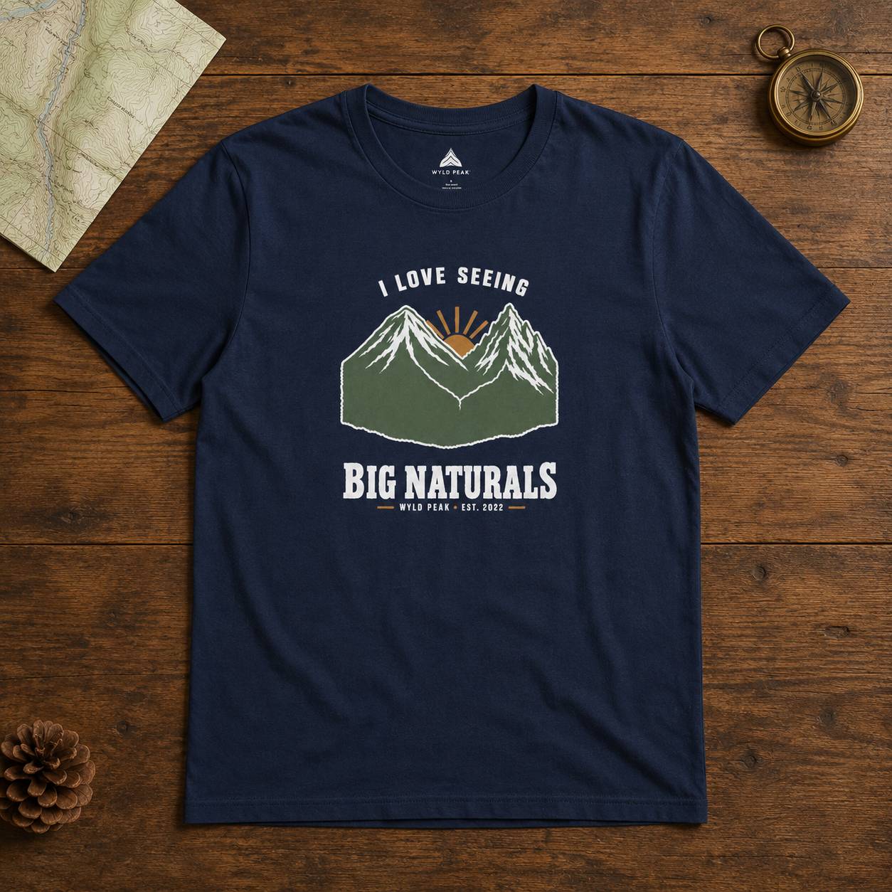 A navy blue graphic t-shirt laid flat on a rustic wooden surface, surrounded by a map, pinecone, and compass. The t-shirt features a graphic of mountains with a sunrise and the text 'I LOVE SEEING BIG NATURALS' and 'WILD LIFE.