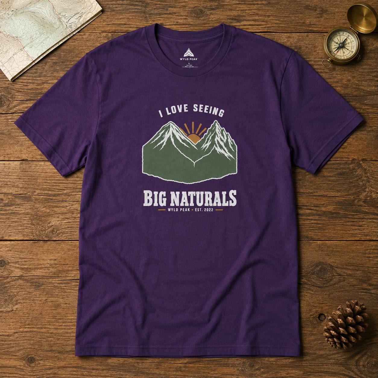 A purple short-sleeve graphic t-shirt laid flat on a rustic wooden surface, surrounded by a map, a vintage compass, and a pinecone. The t-shirt features a graphic of green mountains with a sunburst and the text 'I LOVE SEEING BIG NATURALS WYLD PEAKS EST.