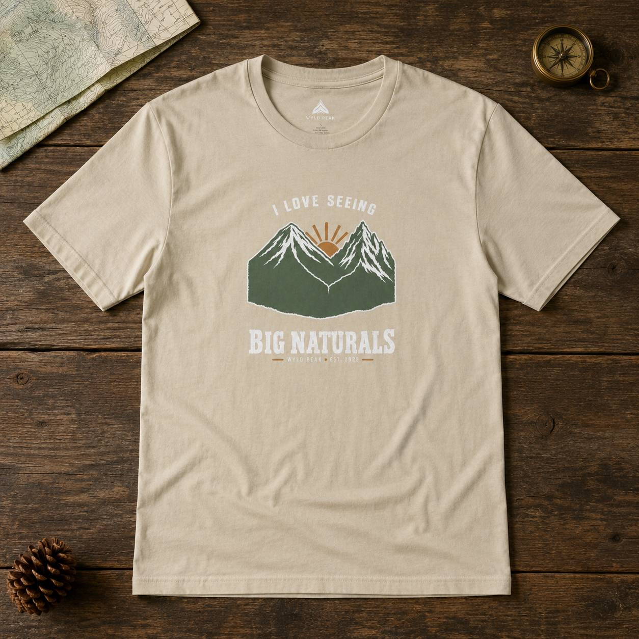 A beige graphic t-shirt laid flat on a rustic dark wood background, surrounded by a map, pinecone, and compass. The t-shirt features a graphic of green mountains with a sunburst and the text 'I LOVE SEEING BIG NATURALS'.