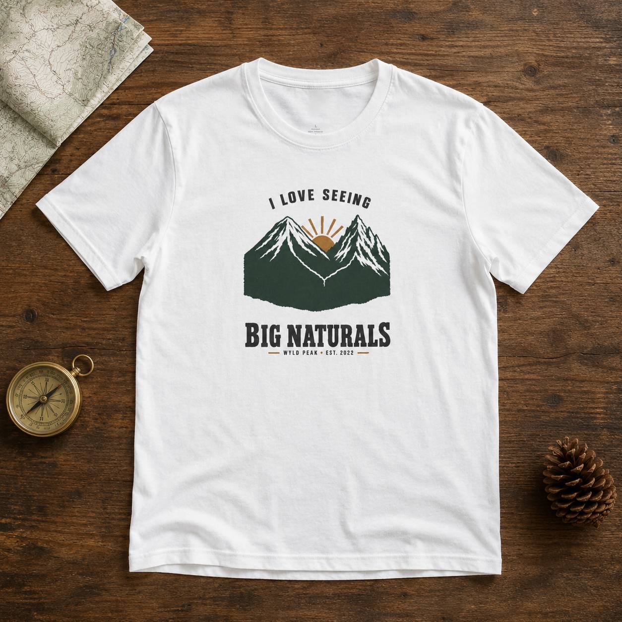 A white short-sleeve graphic T-shirt laid flat on a rustic wooden surface, surrounded by a map, compass, and pinecone. The shirt features a mountain graphic with a sun and the text 'I LOVE SEEING BIG NATURALS WYLD PEAK EST.