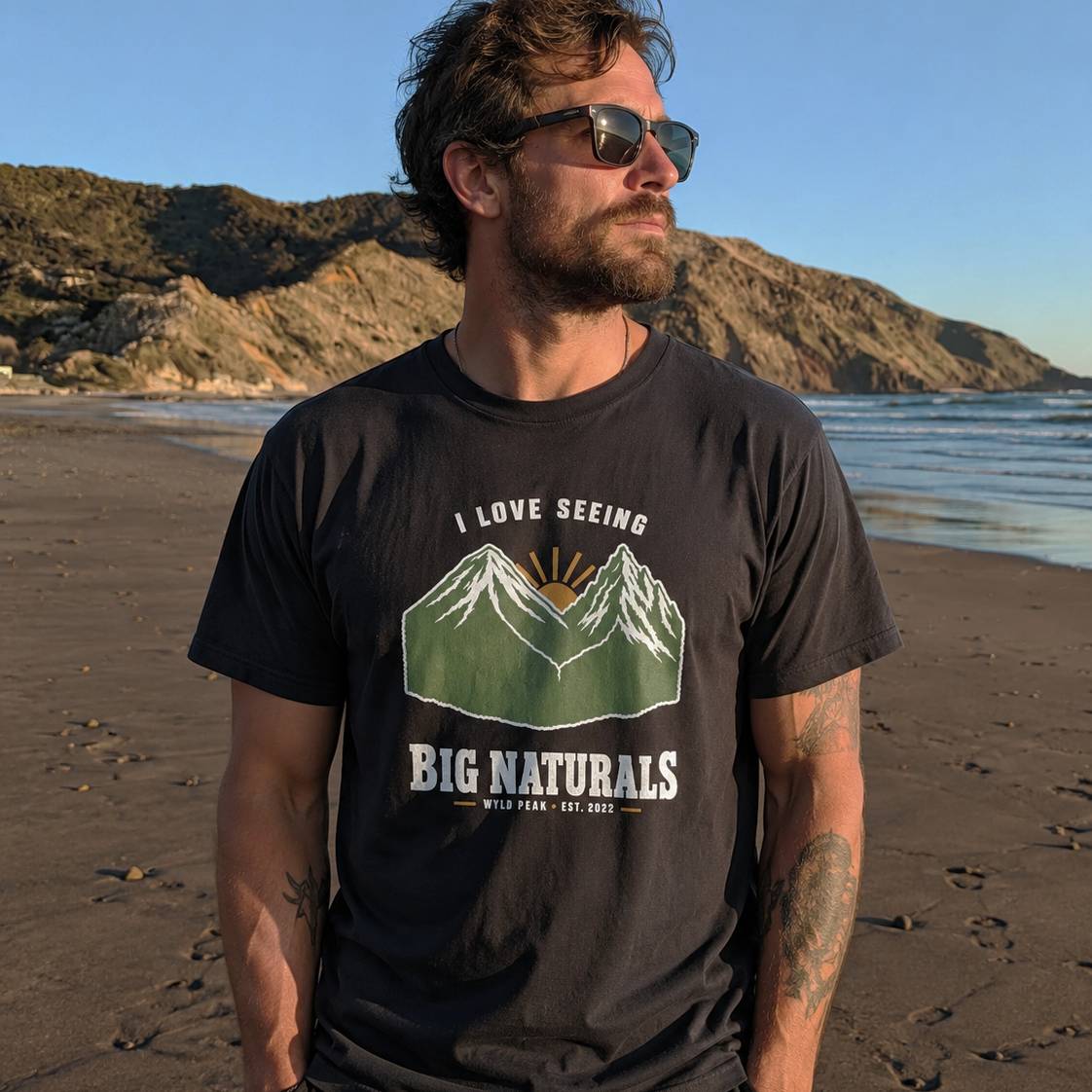 A man wearing a black graphic t-shirt with a mountain design and the text 'I LOVE SEEING BIG NATURALS' stands on a beach with mountains and ocean in the background.