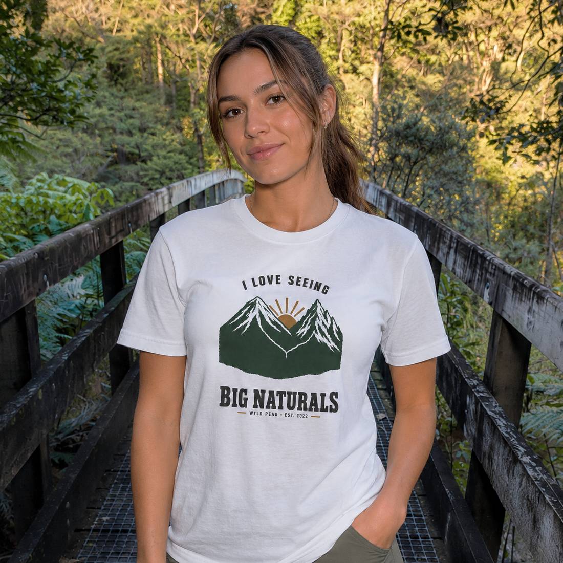 A woman stands on a wooden boardwalk in a forest wearing a white graphic t-shirt with a green mountain and sun design and the text 'I LOVE SEEING BIG NATURALS'.