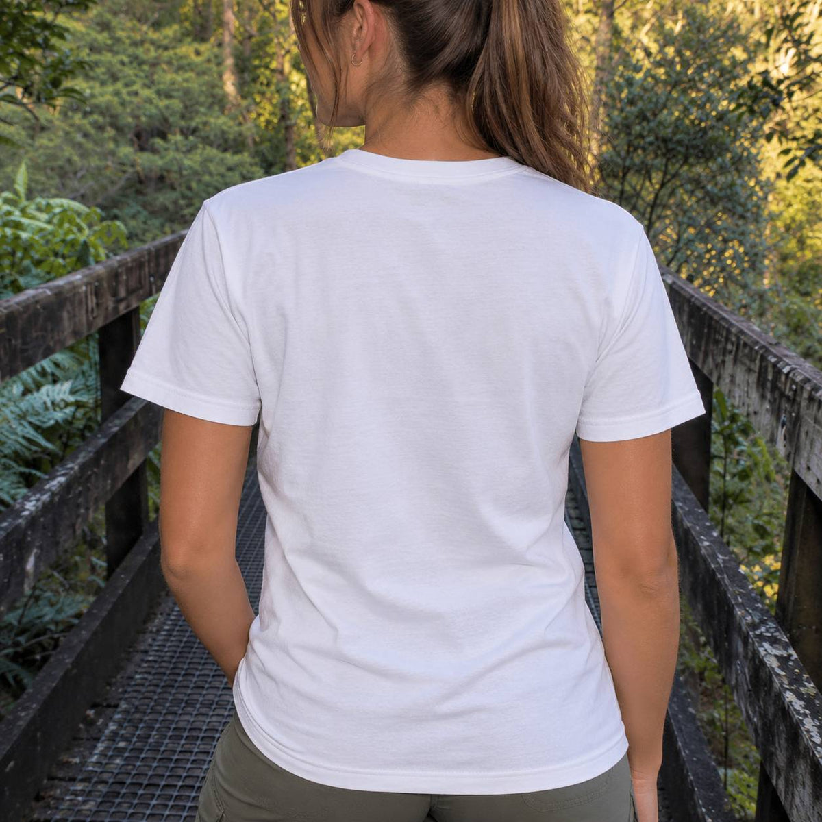 A woman viewed from behind wearing a plain white short-sleeve crewneck t-shirt, standing on a wooden bridge with a lush green forest background.