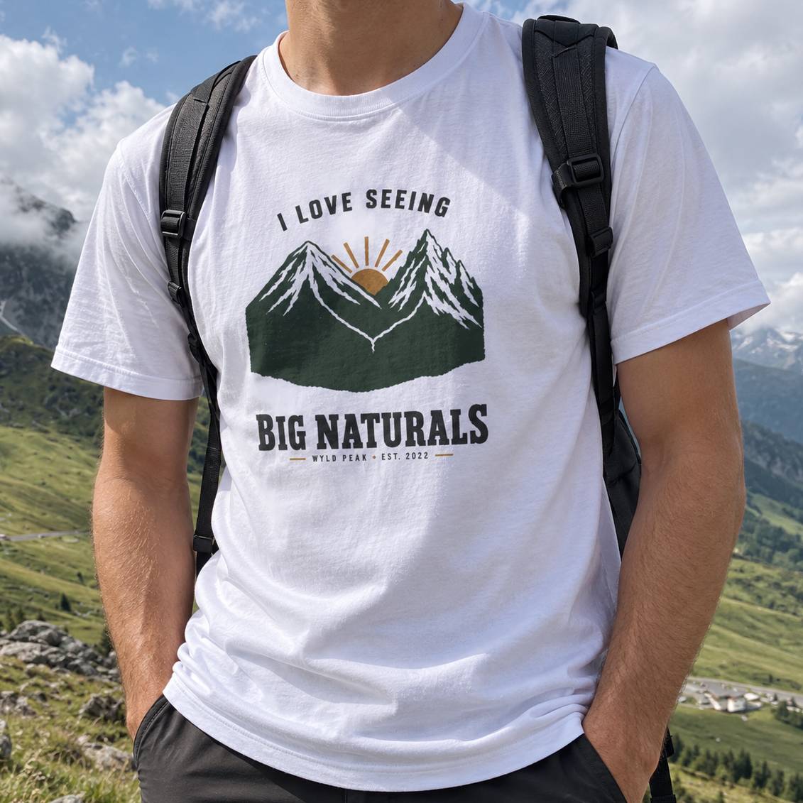 A man wearing a white graphic t-shirt with a mountain and sun design that reads 'I LOVE SEEING BIG NATURALS'. He is outdoors in a mountain landscape with a black backpack on.