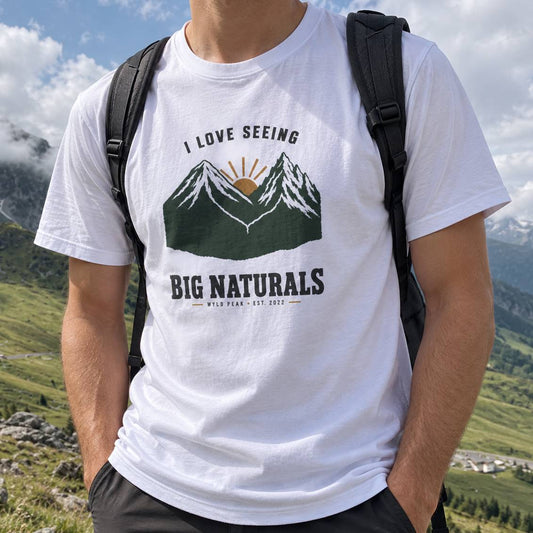 A man wearing a white graphic t-shirt with a mountain and sun design that reads 'I LOVE SEEING BIG NATURALS'. He is outdoors in a mountain landscape with a black backpack on.