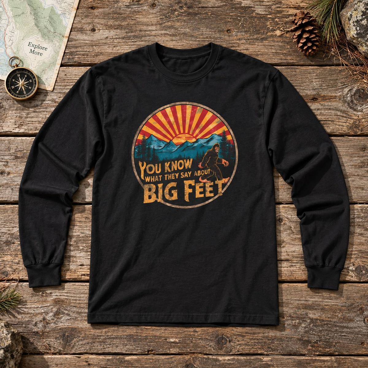A black long-sleeve t-shirt with a retro circular graphic on the chest, laid flat on a rustic wooden surface. The graphic features a sunset over mountains with the text 'YOU KNOW WHAT THEY SAY ABOUT BIG FEET'.
