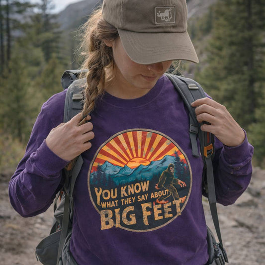 Person wearing a purple sweatshirt with a graphic design and text, standing in a forested area.