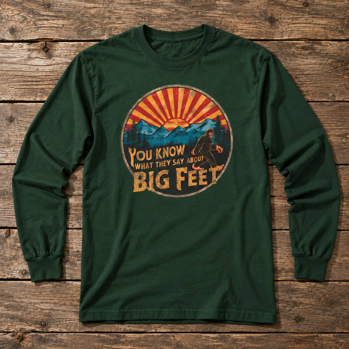 A dark green long-sleeve t-shirt with a circular graphic print on the chest, laid flat on a rustic wooden plank background. The graphic features a sunset over mountains with a Bigfoot silhouette and the text 'YOU KNOW WHAT THEY SAY ABOUT BIG FEET'.