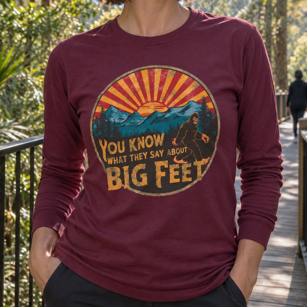 A person wearing a maroon long-sleeve t-shirt with a retro-style Bigfoot graphic on the chest, standing on a wooden boardwalk outdoors.