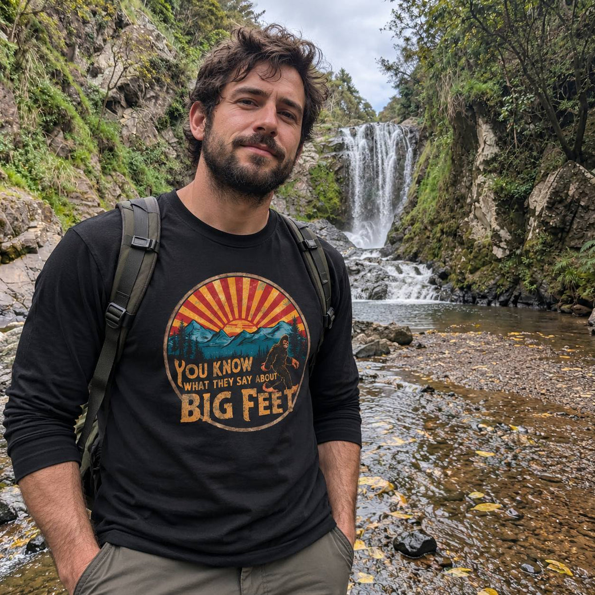 A man wearing a black graphic t-shirt with a 'Big Feet' design, standing outdoors in front of a waterfall in a rocky, mossy mountain stream.