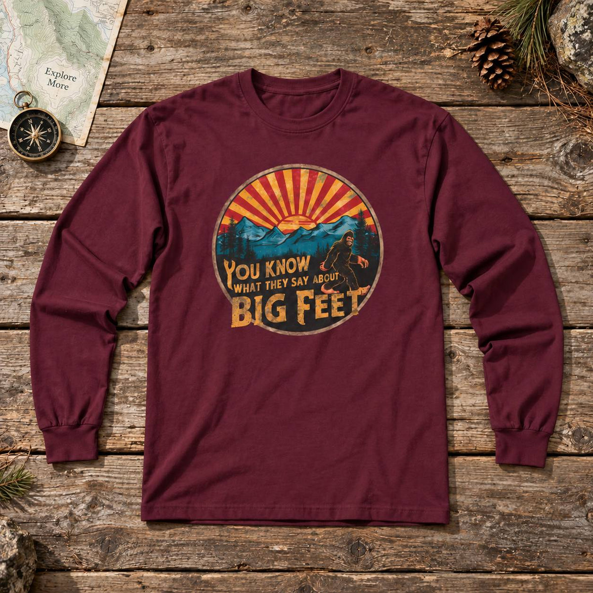 A long-sleeve crewneck t-shirt in maroon, laid flat on a rustic wooden surface. The shirt features a circular graphic print in the center with a sunset over mountains and the text 'YOU KNOW WHAT THEY SAY ABOUT BIG FEET'.