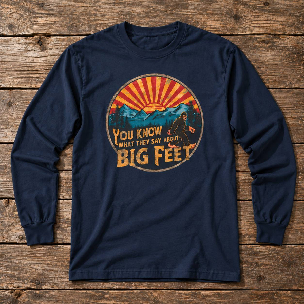 A navy blue long-sleeve t-shirt with a circular graphic print on the chest, laid flat on a rustic wooden plank background. The graphic features a sunset over mountains with Bigfoot in the center and the text 'YOU KNOW WHAT THEY SAY ABOUT BIG FEET'.