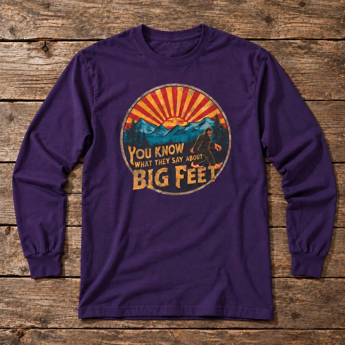 A purple long-sleeve t-shirt with a retro circular graphic on the chest, laid flat on a rustic wooden plank background. The graphic features a mountain sunset with the text 'YOU KNOW WHAT THEY SAY ABOUT BIG FEET'.