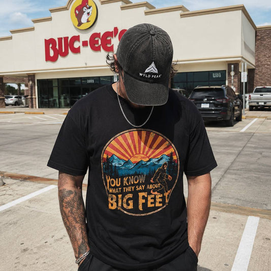 Person wearing a black t-shirt with a graphic design and text, standing in front of a Buc-ee's store.