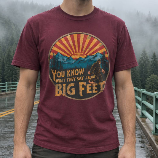 A man wearing a maroon crewneck t-shirt with a vintage-style Bigfoot graphic on the chest, standing on a wet bridge with a misty forest background.