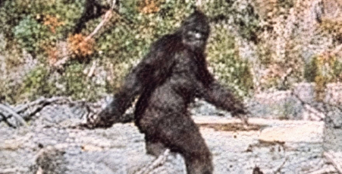 A low-resolution, grainy image depicting a Bigfoot-like creature standing in a forested, rocky outdoor environment.