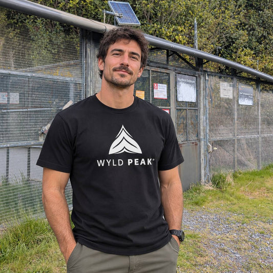 A man wearing a black t-shirt with a white 'WYLD PEAK' logo stands outdoors in front of a chain-link fence and greenery.